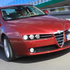 Czerwone Alfa Romeo 159 2.4 JTDm pędzi po drodze, kierowca skupiony na trasie.
