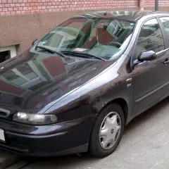 Ciemny Fiat Marea stoi zaparkowany przy ceglanym budynku.