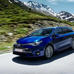 Niebieska Toyota Avensis T27 1.8 benzyna przemierza górską drogę, otoczona zielonymi zboczami i ośnieżonymi szczytami.