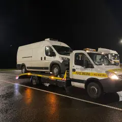Nocne holowanie na drodze ekspresowej. Biała furgonetka jest transportowana na lawecie pomocy drogowej.