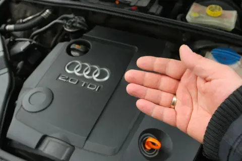 Dłoń pokazuje silnik Audi 2.0 TDI. Widoczny jest pomarańczowy bagnet do sprawdzania poziomu oleju, a obok niego korek, pod którym może znajdować się masło pod korkiem oleju.