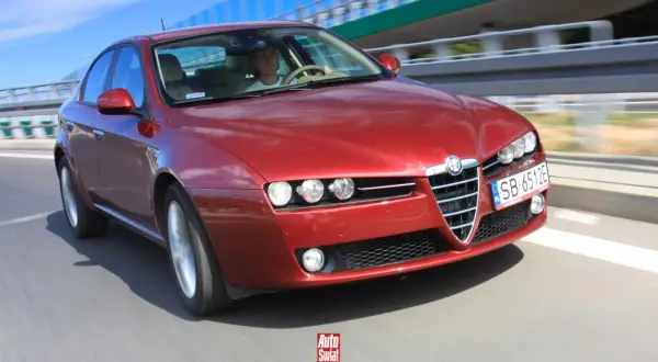 Czerwone Alfa Romeo 159 1.9 JTD pędzi po drodze. Kierowca w białej koszulce pewnie trzyma kierownicę.