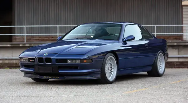 Granatowy Alpina B12 z lat 90. błyszczy na tle industrialnego budynku. Złote detale podkreślają jego sportowy charakter.