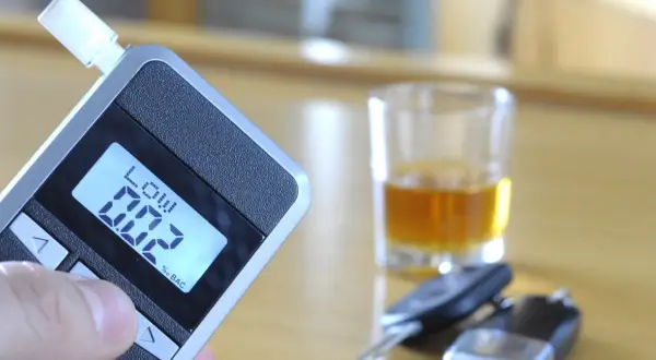 Alkomat z wyświetlaczem 0.02% BAC, szklanka z alkoholem i kluczyki samochodowe.