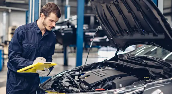 Mechanik w granatowym kombinezonie sprawdza listę kontrolną przy otwartej masce samochodu. Konie mechaniczne skrót do szybkiej diagnozy.