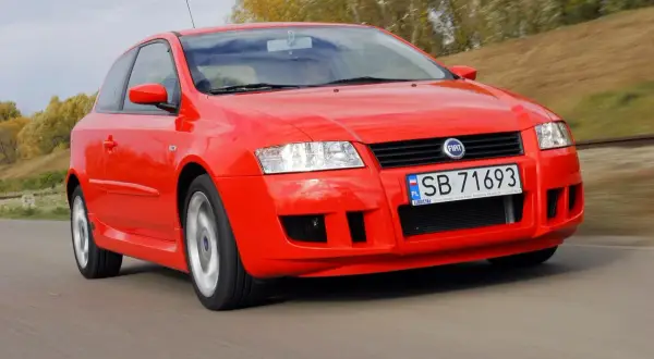 Czerwony Fiat Stilo 1.6 16v pędzi po drodze, otoczony jesienną zielenią.