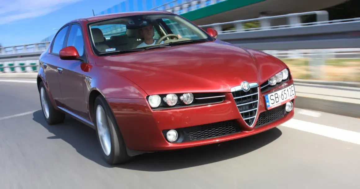 Czerwone Alfa Romeo 159 2.4 JTDm pędzi po drodze, kierowca skupiony na trasie.