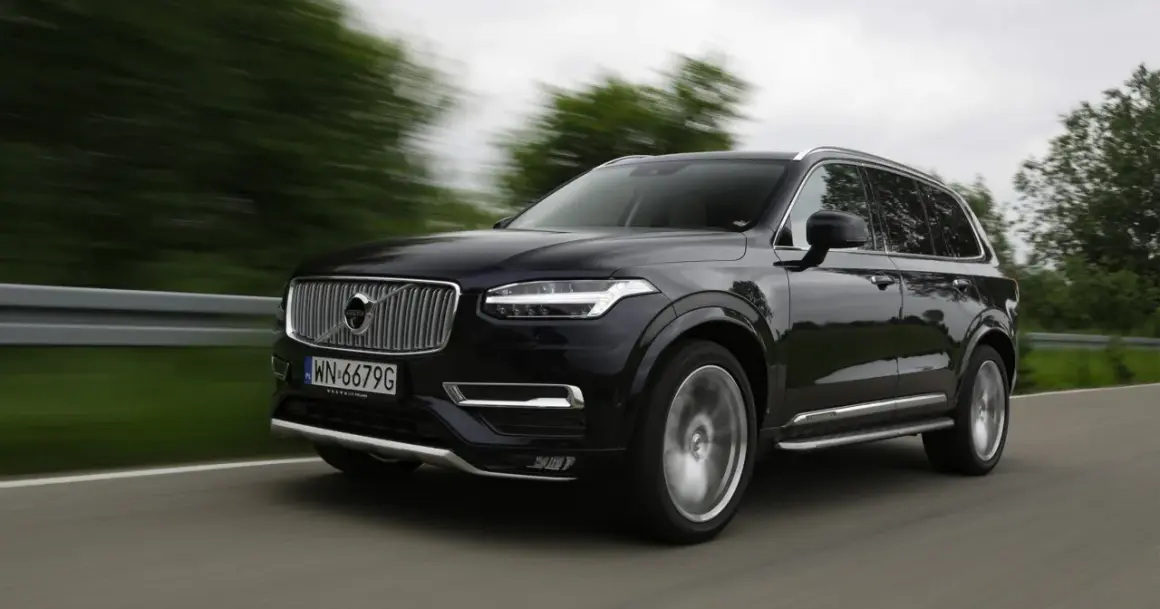 Czarne Volvo XC90 V8 pędzi po drodze, rozmazane drzewa i krajobraz świadczą o dużej prędkości.