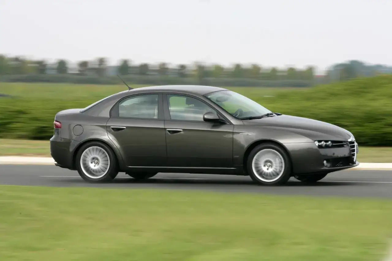 Szary Alfa Romeo 159 2.4 JTDm pędzi po drodze, otoczony rozmytą zielenią.