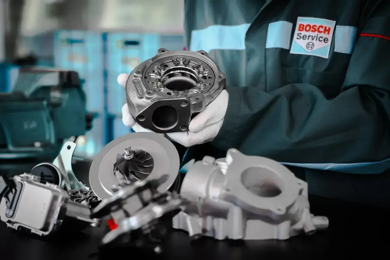Mechanik w rękawiczkach prezentuje części turbosprężarki, sugerując domowe ustawienie sztangi turbiny.