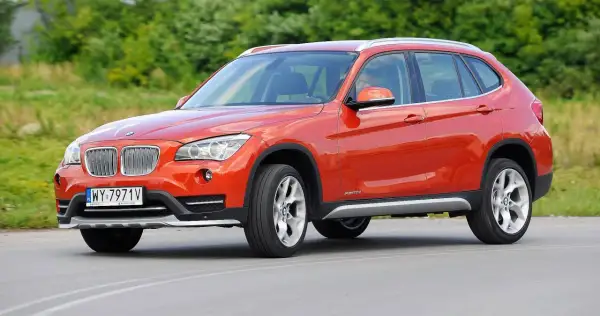 Pomarańczowe BMW E84 X1 z polskimi tablicami rejestracyjnymi jedzie po drodze, otoczone zielenią.