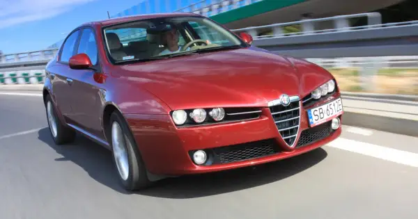 Czerwone Alfa Romeo 159 2.4 JTDm pędzi po drodze, kierowca skupiony na trasie.