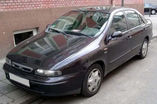 Ciemny Fiat Marea stoi zaparkowany przy ceglanym budynku.