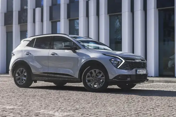 Nowy Kia Sportage Anniversary w jasnym kolorze, zaparkowany przed nowoczesnym budynkiem. Stylowy SUV na kamienistej nawierzchni.