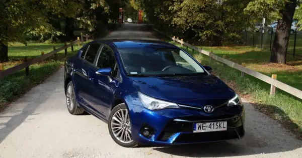 Błękitna Toyota Avensis T27 na żwirowej drodze, otoczona zielenią i drzewami.
