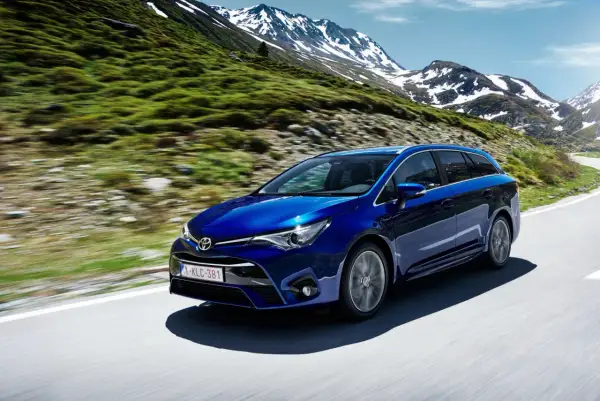 Niebieska Toyota Avensis T27 1.8 benzyna przemierza górską drogę, otoczona zielonymi zboczami i ośnieżonymi szczytami.