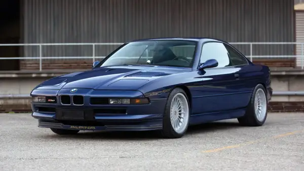 Granatowy Alpina B12 z lat 90. błyszczy na tle industrialnego budynku. Złote detale podkreślają jego sportowy charakter.