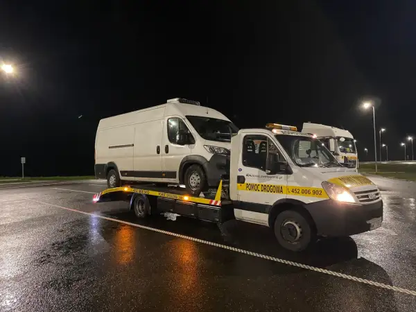 Nocne holowanie na drodze ekspresowej. Biała furgonetka jest transportowana na lawecie pomocy drogowej.