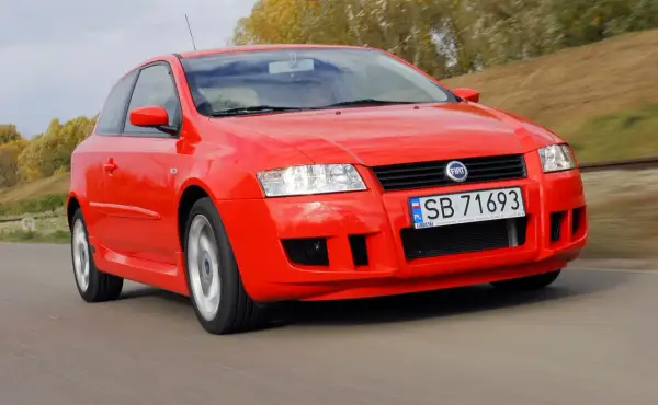 Czerwony Fiat Stilo 1.6 16v pędzi po drodze, otoczony jesienną zielenią.