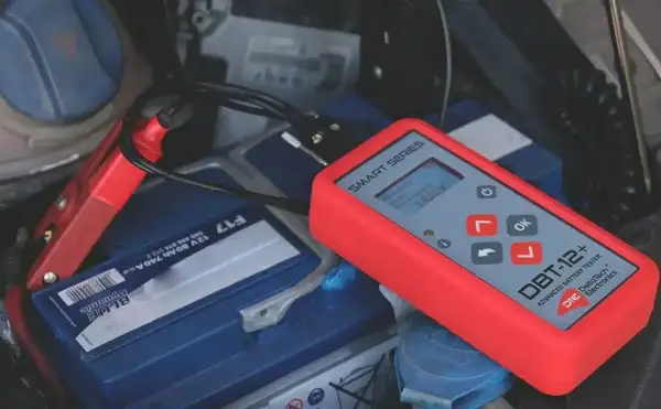 Czerwony tester akumulatorów podłączony do akumulatora samochodowego. Wyświetla 14.25V, sugerując, że problemem nie jest rozładowany akumulator, a coś innego.
