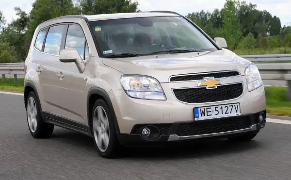 Złoty Chevrolet Orlando 1.8 benzyna na drodze. Rodzinny van w ruchu, z widocznym kierowcą.