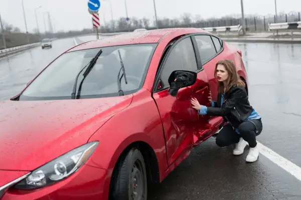 Młoda kobieta ogląda uszkodzone drzwi czerwonego samochodu po wypadku. Widać wgniecenie i urwane lusterko. Zawieszenie auta mogło ucierpieć.